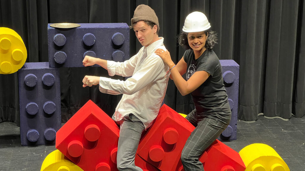 Zwei Personen auf übergroßen Legosteinen. Vorn sitzt ein Mann, hinten eine Frau. Beide befinden sich auf einer Bühne oder auf einem Bühnenaufbau bei den Proben zum neu inszenierten Theaterstück "Aschenputtel"