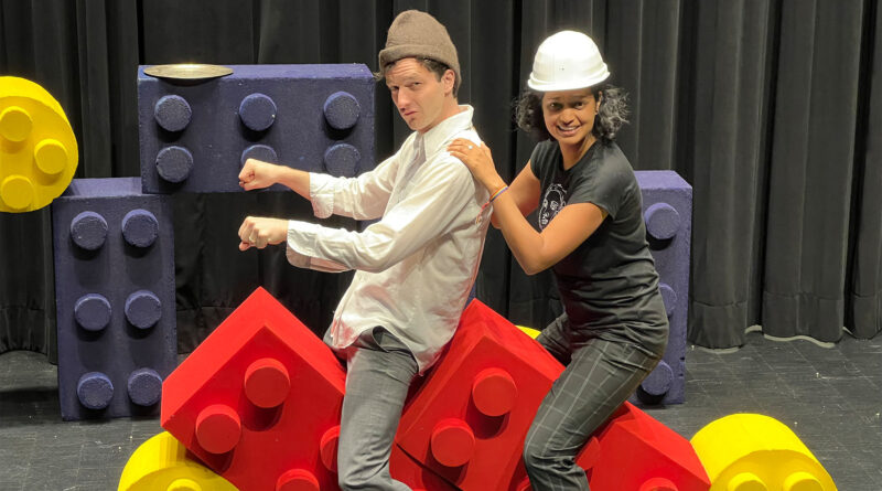 Zwei Personen auf übergroßen Legosteinen. Vorn sitzt ein Mann, hinten eine Frau. Beide befinden sich auf einer Bühne oder auf einem Bühnenaufbau bei den Proben zum neu inszenierten Theaterstück "Aschenputtel"