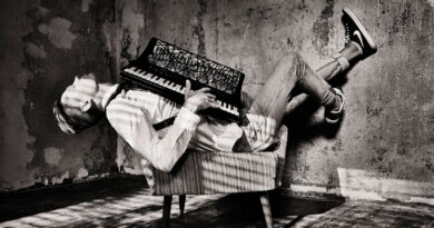 Schwarzweißfoto eines Mannes, der entspannt in einem Sessel liegt und ein Akkordeon auf der Brust hält, in einem rauen, industriell wirkenden Raum.