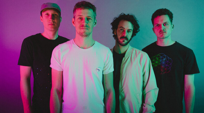 Vier Männer stehen nebeneinander vor einem farbig beleuchteten Hintergrund in Rosa und Türkis. Sie tragen legere Kleidung und schauen direkt in die Kamera; das Licht erzeugt einen neonartigen Effekt auf dem Bild.